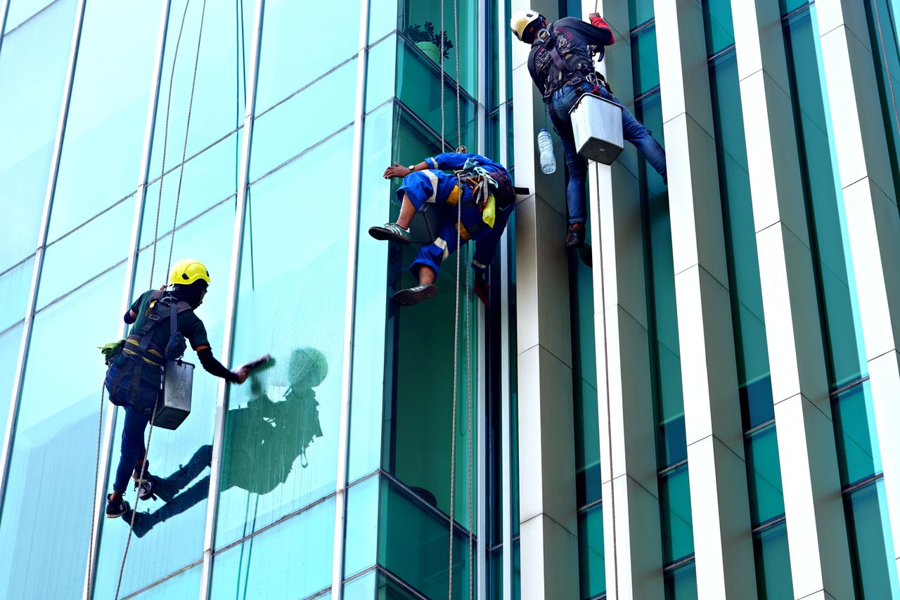 services-02 Three window cleaners suspended on ropes clean a modern glass building facade during the day.